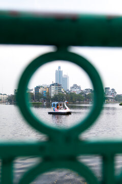 View of a swan paddle boat floats serenely on the water framed by a green metal railing, city buildings rise in the background, Hanoi, Ha Noi, Vietnam.