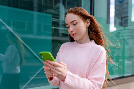 Young woman checks phone while standing near building with glass windows in a city setting