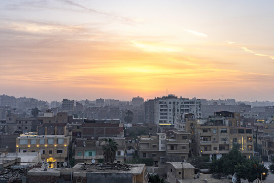 Aerial view of dense urban sprawl under a warm, glowing sky, the buildings a mix of textures and heights, casting shadows in the soft light, Giza, Cairo, Egypt.