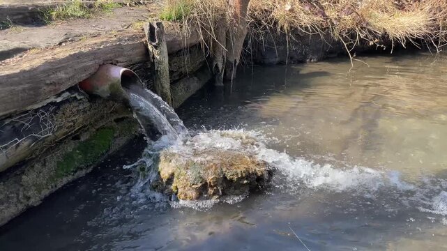 Rusty pipe discharges water into creek, mosscovered outlet splashes foam against wooden bank, churning sediment and algae under cold winter