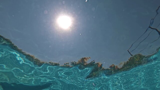Underwater view of a splash with bubbles beneath a sunlit pool surface against blue sky and clouds. Concept for summer, recreation, wellness, sport, travel, and water background design.