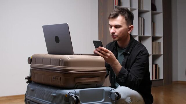 Man uses stacked suitcases as a makeshift desk, working on a laptop and checking his smartphone while preparing for a nomadic remote work lifestyle and upcoming trip