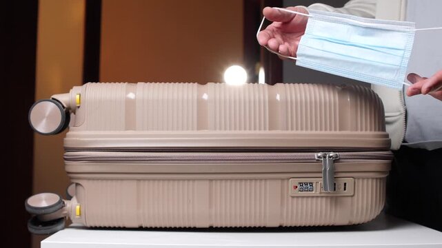 Person's hands holding a blue medical face mask above a beige hard shell suitcase, preparing for a trip while prioritizing health and safety during the journey