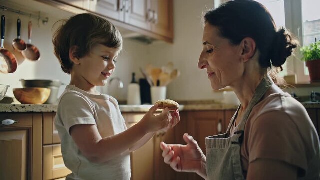 Child with brown hair offers a cookie to an older woman in a kitchen, both smiling and engaged in a warm interaction, with wooden cabinets and utensils visible
