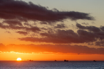Fototapeta premium Sunset over the Atlantic ocean with freight ships passing near the descending sun, near the port of Huelva, Spain