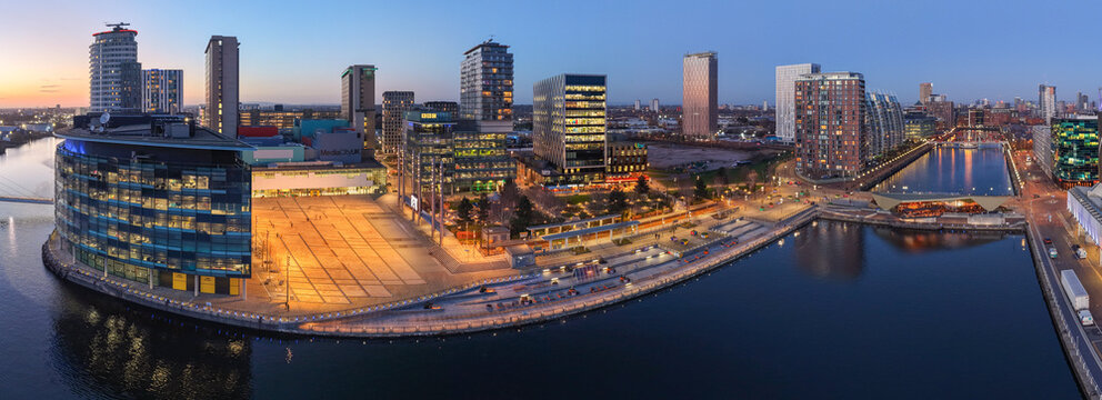 A wide aerial panoramic view of the MediaCityUK development in Salford Quays, featuring the BBC North buildings, ITV studios, and the central plaza reflected in the Manchester Ship Canal at dusk.
