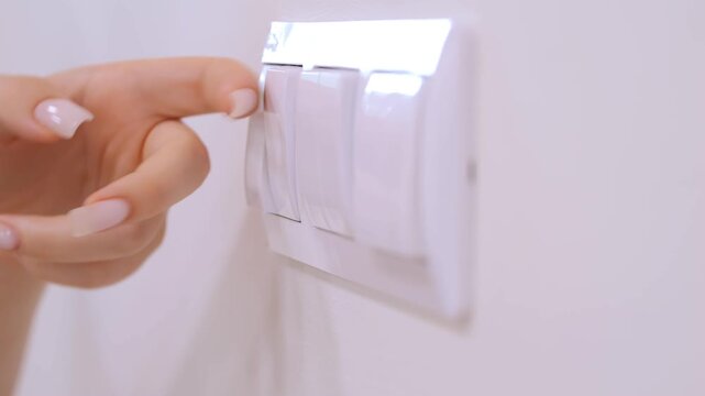 Hand with manicured nails presses wall switch. White switch with three toggles. Wall is plain and light-colored. Smooth motion from pressing to releasing switch
