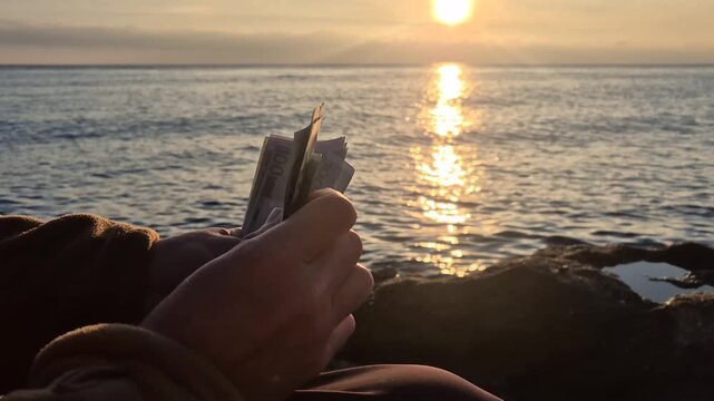 Money counting sunset, hands tallying 100 euro notes while sitting by the ocean water at glorious golden hour
