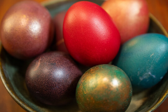 a brown ceramic bowl filled with dyed easter eggs in shades of vibrant red, deep purple, and metallic green-bronze, resting on a dark wooden surface under warm natural light.
