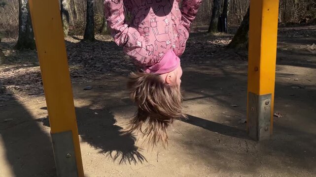 Toddler girl hanging upside down from a playground bar outdoors, enjoying active play and developing balance and strength.