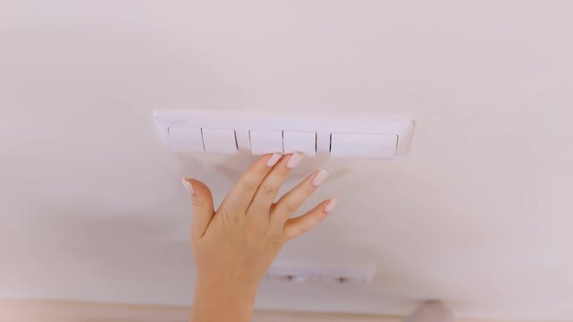 Hand reaches up to touch light switch. Fingers hover above white wall-mounted switch. Soft lighting illuminates scene from above. Focus on hand and switch only. No animals or plants visible