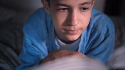 Obraz premium Thoughtful teenage boy reading a book in dim light, have fun and enjoy