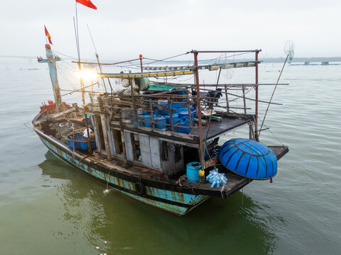 Aerial view of a weathered fishing boat with faded blue paint and a red flag bobbing gently on the still, grey waters, Da Nang, Vietnam.