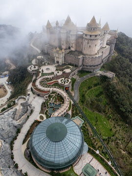 Aerial view of a fairytale castle piercing through the mist-laden peaks, with winding pathways below, Ba Na Hills, Vietnam.