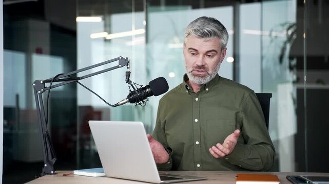 Mature gray-haired businessman recording online podcast in modern office, speaking into microphone using laptop, confident coach conducting virtual business training and sharing professional knowledge