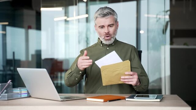 Disappointed gray-haired businessman sitting at office desk reading letter with bad news. Upset mature man worried and frustrated about business failure, loss or financial problem at modern workplace.
