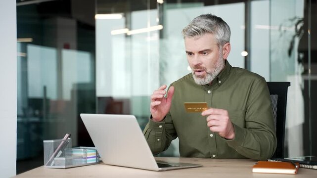 Shocked gray-haired businessman sitting at office desk checking credit card on laptop. Worried mature man discovering online fraud, bank problem, unauthorized payment while working at modern workplace