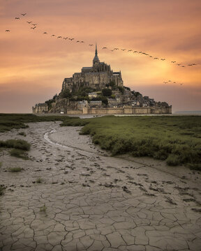 View of Mont Saint-Michel, a medieval abbey perched on a tidal island, rises majestically against a fiery sunset sky with birds flying, Mont Saint Michel, France.