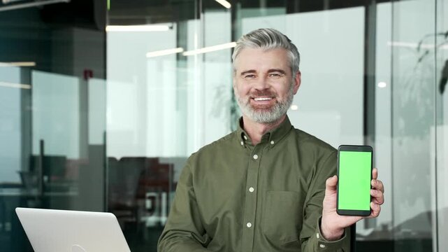 Smiling mature businessman sitting in modern office, looking at camera and showing smartphone with green screen, vertical template layout for advertising, app promotion, digital marketing presentation
