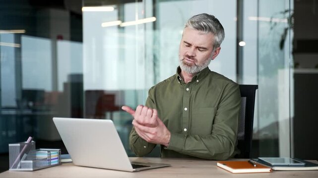 Gray-haired businessman sitting at office desk working on laptop and suffering from wrist pain. Mature employee massaging joint and doing hand exercises to relieve tension and discomfort at workplace.
