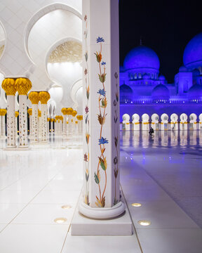 View of ornate white pillars adorned with colorful floral patterns leading to the grand mosque illuminated in blue lights, Abu Dhabi, United Arab Emirates.