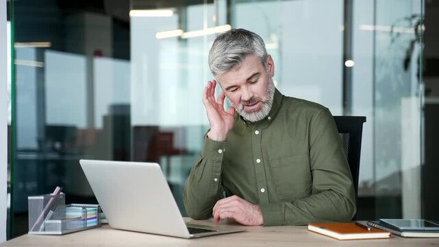 Sick gray-haired businessman sitting at office desk suffering from ear pain. Mature man touching ear with fingers, feeling discomfort and ache caused by infection, otitis or ear disease at workplace.
