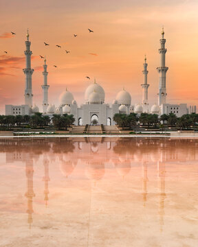 View of the majestic white mosque is mirrored in serene waters under a vibrant sky as birds fly towards the horizon, Abu Dhabi, United Arab Emirates.