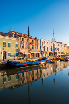 View of a canal reflects a colorful row of buildings and a classic sailing boat in the golden light of Cesenatico, Cesenatico, Italia.