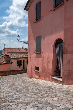 View of the rustic charm of a salmon-colored building with dark shutters, beside a cobblestone street, under a sky dappled with clouds, Longiano, Italia.