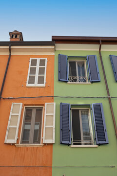 View of a building facade split between vibrant orange and pale green, showcasing contrasting window shutters under a clear sky, Ravenna, Italia.