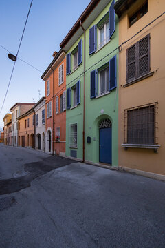 View of vibrant houses in orange, green, and yellow hues stand side-by-side along a quiet, winding asphalt road under a clear sky, Ravenna, Italia.