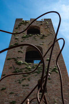 View of a brick tower partially covered with greenery stands tall, framed by an abstract rusty metal sculpture reaching towards the bright blue sky, Longiano, Italia.