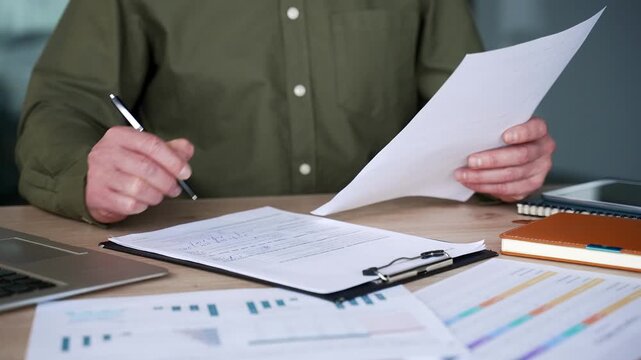 Close-up of businessman hands holding document and writing on clipboard at desk in modern office, filling business form and taking notes, focused professional reviewing paperwork and managing tasks.
