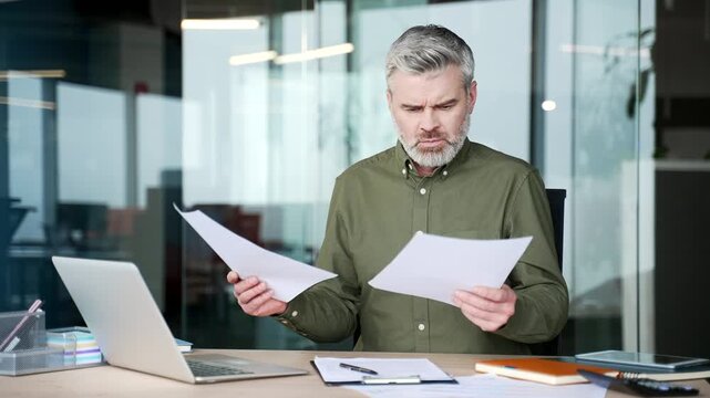 Confused mature businessman sitting at desk in office, looking at documents and at camera with puzzled expression, struggling to understand paperwork, uncertainty and hesitation in business decision.