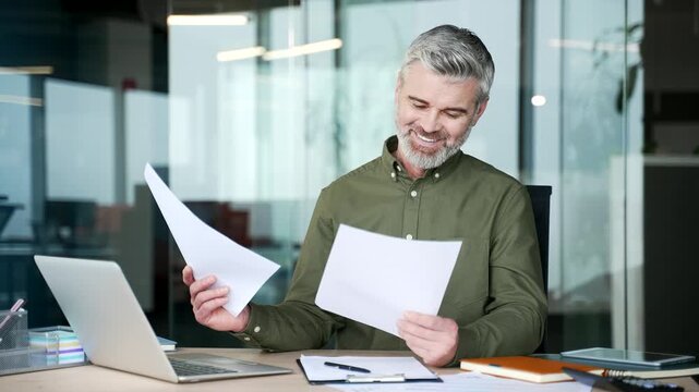 Mature gray-haired businessman sitting at desk in office, holding documents and smiling with satisfaction after reviewing positive business report, feeling proud of successful performance achievements