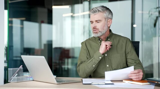 Mature gray-haired businessman sitting at desk in office, holding document and thoughtfully comparing information on laptop screen, focused professional analyzing data and reviewing business report