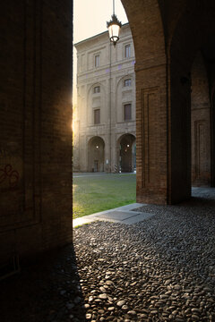 View of sunlight filtering through arched brick corridors onto a cobblestone path leading to a sunlit courtyard with a building, Parma, Emilia Romagna, Italy.