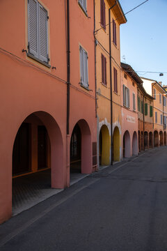 View of sun-drenched buildings with arched walkways cast long shadows on the quiet street, creating a harmonious blend of light and shadow, Parma, Emilia Romagna, Italy.