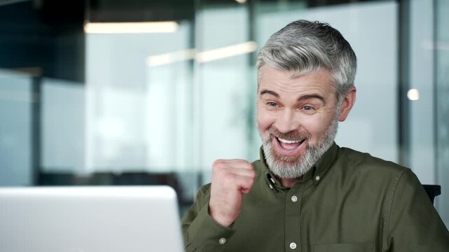 Close-up of gray-haired businessman smiling after reading positive news on laptop. Man reacting with joy and relief while checking good message at workplace in modern office, cheerful and inspired.