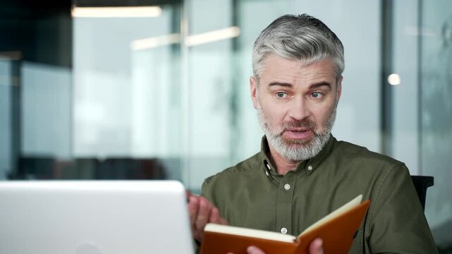 Gray-haired businessman talking, asking questions during video call conference on laptop, taking notes in notebook. Engaging in discussion during remote online meeting at workplace in office. close-up