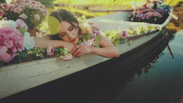 portrait woman goddes sping lying in wooden boat, hand touching stroking surface of water river lake. decor blooming flowers white pink color. Fairy tale elf girl fashion model floating beauty face 4k
