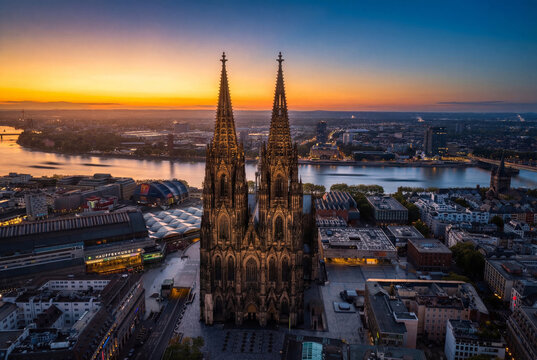 Aerial view of the majestic Cologne Cathedral piercing the skyline at dusk, its gothic spires contrasting with the tranquil Rhine River, Cologne, North Rhine-Westphalia, Germany.