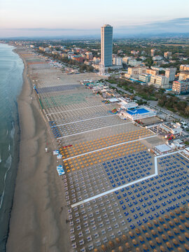 Aerial view of orderly rows of beach umbrellas create a colorful mosaic against the shore, shadowed by a tall building, Sorrivoli, Emilia Romagna, Italia.