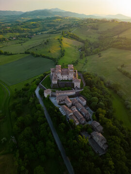 Aerial view of a majestic castle sitting atop a hill, its stone walls glowing softly against the backdrop of green fields, Parma, Emilia Romagna, Italy.