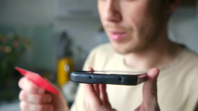 Naive man holding bank card and smartphone, dictating his financial data to scammer during phone call at home, becoming victim of online fraud, identity theft. Concept of phone fraud or scammer.