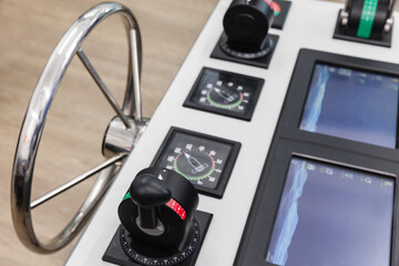 Close-up of a ships bridge helm showing a stainless steel steering wheel, throttle controls, gauges and navigation displays conveying marine control, precision steering and modern nautical technology. © evannovostro