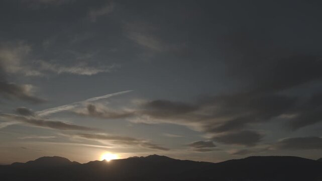 Dawn breaks behind the silhouette of a mountain, with the white contrail of an airplane streaking across the sky. Dark clouds and golden light create a cinematic landscape. A concept of travel and new