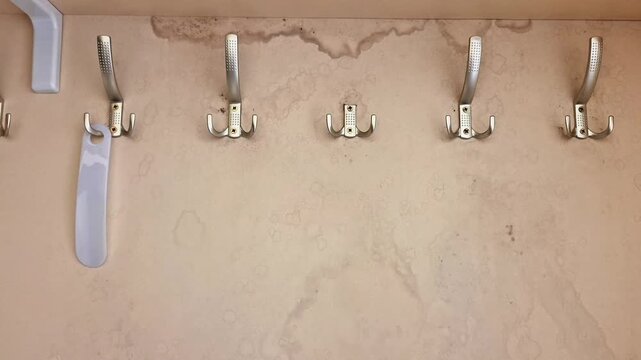 Row of six metallic wall hooks mounted on a beige textured wall, one hook holding a white plastic item, with visible staining above showing signs of water damage