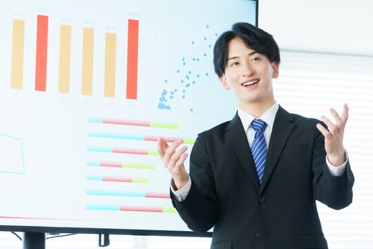 オフィスでディスプレイに映ったグラフを説明しながらプレゼンをする若い日本人男性