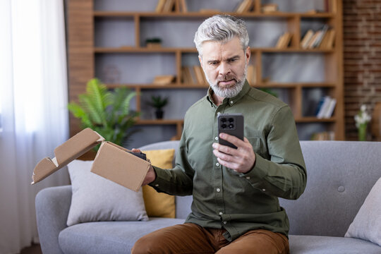 Mature man holding an open cardboard box and smartphone, looking disappointed and stressed after receiving an incorrect or damaged online order at home